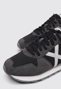 Zapatillas negras y grises que combinan materiales de ante y malla, con un detalle en cruz blanco, punta redonda y cordones texturizados.