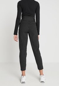 Woman standing facing away wearing black long-sleeve top, black pinstriped pants, and white sneakers on plain background.