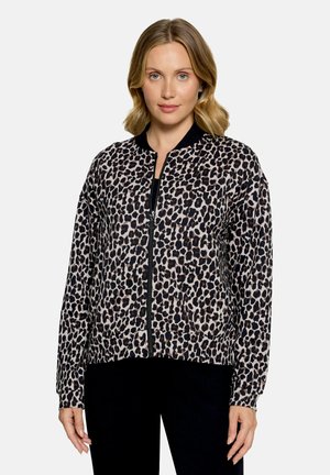 Femme portant une veste zippée imprimé léopard avec col et poignets noirs, se tenant devant un fond clair uni.