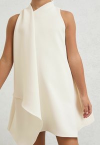 Fehér, ujjatlan ruha aszimmetrikus redőzéssel. Sima anyag, magas nyak és légiesen folyó alj.