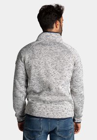 Man in een lichtgrijze gemêleerde ritstrui en blauwe jeans, gezien van achteren tegen een effen achtergrond.