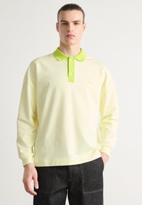 Camisa de polo amarilla clara de manga larga con un cuello y botones de un verde brillante en contraste; presenta un pequeño logotipo bordado en el pecho.