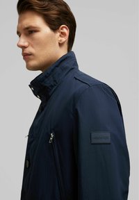Navy windbreaker jacket, lichte stof. Kenmerken zijn een hoge kraag, drukknoopsluiting en een ritszak. Merkpunt op de mouw.
