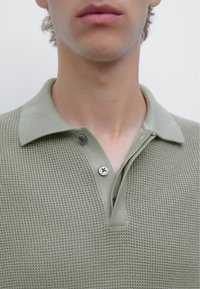 Polo shirt lavorato a maglia verde chiaro con un tessuto texturizzato,patta con tre bottoni e un design del colletto classico. Ingrandimento della parte superiore del capo.