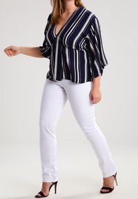 Donna che indossa una blusa a righe blu navy, pantaloni bianchi e sandali neri con tacco alto, in piedi davanti a uno sfondo chiaro e uniforme.