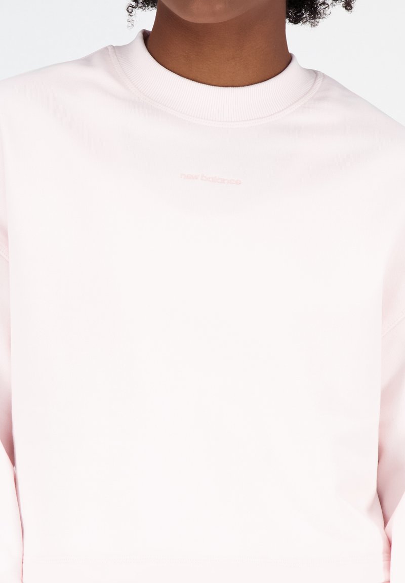 Sudadera de cuello redondo de color rosa claro, utilizada por una persona con un pequeño logotipo de "new balance" bordado en el pecho.