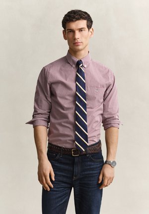 Homme portant une chemise à carreaux retroussée, une cravate rayée, un jean foncé et une montre, debout devant un fond clair uni.