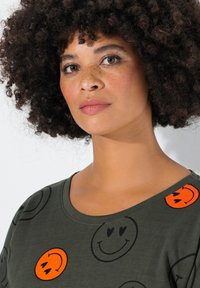Camiseta verde oliva con cuello redondeado, con patrones de caras sonrientes en negro y naranja. Fabricada en un tejido suave con un corte casual.