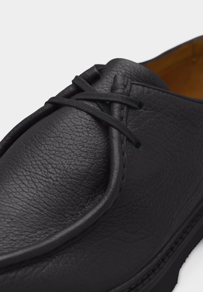Mocassin en cuir noir texturé avec un bout arrondi, une couture style mocassin et un lacet en cuir noir noué simplement.