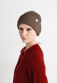 Bonnet en tricot côtelé marron avec un revers, arborant une petite étiquette de logo blanche. Le modèle porte un pull rouge, avec un fond neutre.
