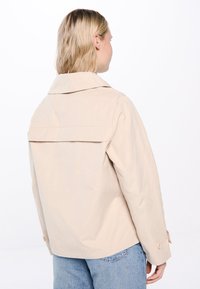 Beige takki, jossa on leveä kaulus ja takana yksityiskohta. Valmistettu kevyestä materiaalista, jossa on hihnapainikkeet ja rento siluetti.