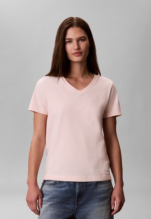 Γυναίκα με ίσια καστανά μαλλιά που φοράει ένα ανοιχτό ροζ μπλουζάκι με V-λαιμόκοψη και κοντά μανίκια και μπλε τζιν σε απλό γκρι φόντο.