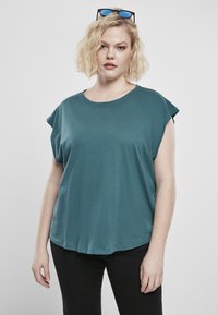 Tealfärgad T-shirt med korta ärmar, rund halsringning och uppvikta muddar. Mjuk textur med en lös passform, bärs tillsammans med svarta byxor.