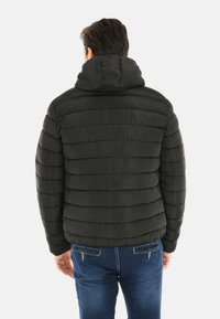 Uomo che indossa una giacca a vento nera con cappuccio e jeans blu, in piedi con la schiena rivolta verso uno sfondo bianco semplice.