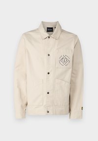 Beige jacket made of cotton with a collar, front pockets, and a logo patch on the chest. Features button closure and long sleeves.