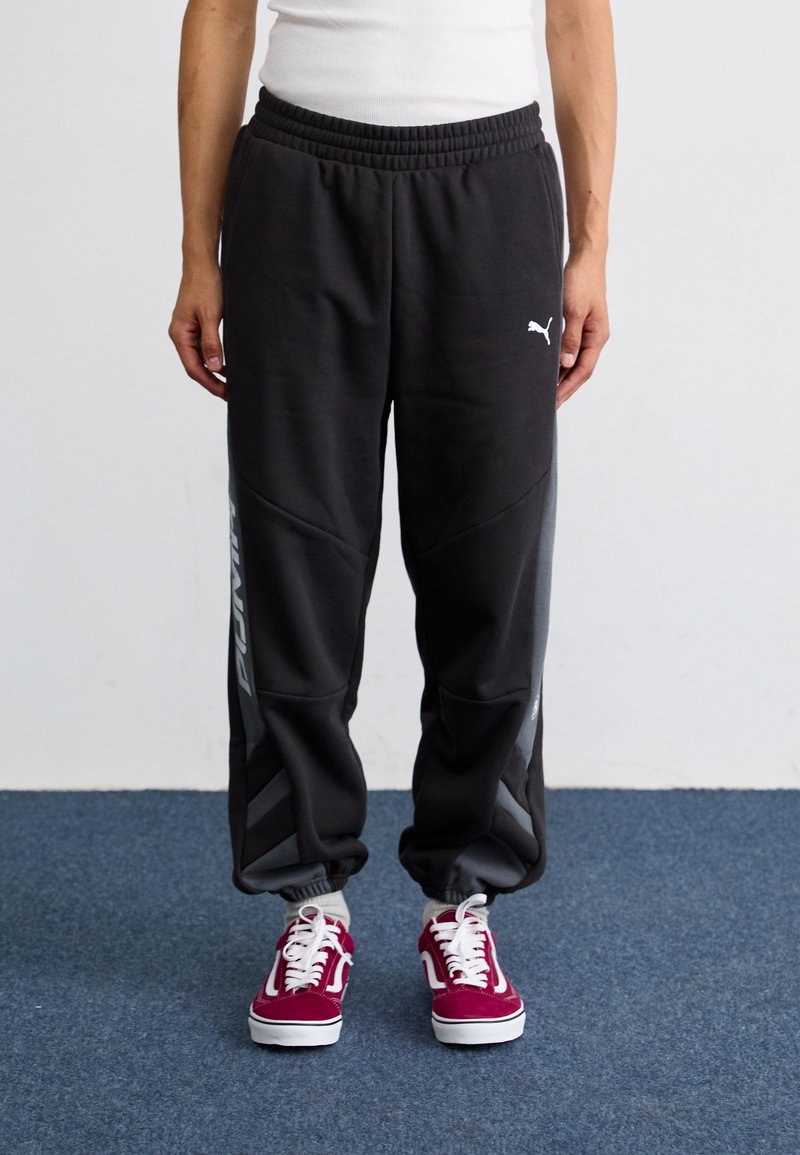 Fekete melegítónadrág elasztikus derékkal, oldalsó zsebekkel és szürke részletekkel. Puma logóval és szűkített mandzsettával rendelkezik, burgundi színű sportcipővel párosítva.