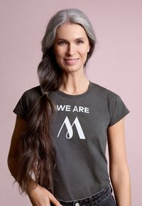 Dark grey T-shirt with white text "WE ARE" and a stylized "M" design, short sleeves, soft texture, paired with black high-waisted jeans.