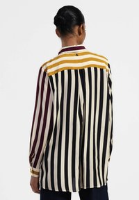 Donna con capelli scuri raccolti in uno chignon, che indossa una camicia a maniche lunghe con strisce verticali nere, bianche, amaranto e gialle, vista da dietro.