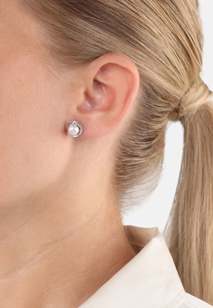 Pendientes de plata con un centro de perla redonda, acentuados con una pequeña gema transparente, mostrados en la oreja de una persona.