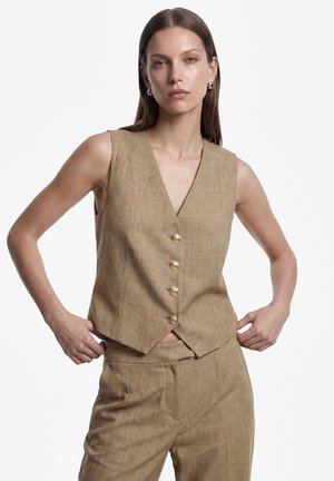 Kvinde med glat brunt hår iført en ærmeløs beige vest med guldknapper og matchende beige bukser, stående med hænderne nær taljen.
