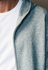 Grijze zip-up hoodie gemaakt van zacht, gestructureerd materiaal, met een close-up van de kraag en een wit t-shirt met ronde hals eronder.