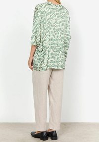 Blouse ample à motif abstrait vert et crème, associée à un pantalon beige clair. Chaussures noires à bouts carrés complètent la tenue.