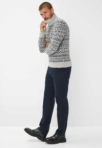 Maglione a collo alto a maglia grigia con motivo, rifinito con dettagli in blu navy, abbinato a pantaloni blu navy scuri e scarpe stringate nere su uno sfondo semplice.