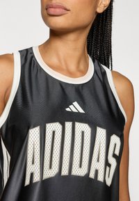 Juoda atletinė liemenė su balta briauna, dideliu kremine spalva parašytu „ADIDAS“ tekstu. Medžiaga atrodo lygi ir drėgmę sugerianti.