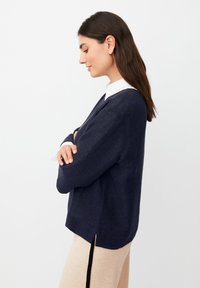 Maglione a maglia blu navy con scollo a V, maniche lunghe e spacchi laterali. Indossato su una camicia leggera con colletto, con una finitura testurizzata.