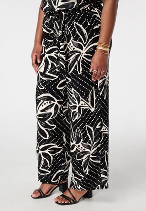 Zwarte broek met wijde pijpen en grote witte bloemenprint, gecombineerd met zwarte sandalen met bandjes en gouden armbanden aan de rechterpols.