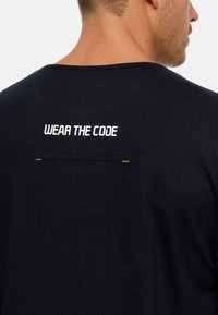 Camiseta de algodón negra con cuello redondo; presenta el texto blanco "LLEVA EL CÓDIGO" bordado en la parte superior de la espalda, con costuras de acento en naranja.