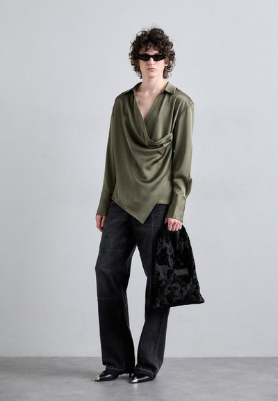 Langärmlige, olivgrüne, drapierte Bluse mit Kragen, kombiniert mit dunklen, weit geschnittenen Jeans und einer schwarzen, strukturierten Tote-Tasche. Spitze Schuhe mit silberner Zehenkappe.