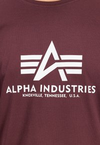 Bordeauxfarbener Baumwoll-T-Shirt mit einem großen weißen Grafiklogo, das den Text "ALPHA INDUSTRIES" und "KNOXVILLE, TENNESSEE, U.S.A." darunter zeigt.