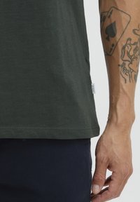 Camiseta de algodón de color verde oscuro con cuello redondo, mangas cortas y una textura sutil. Presenta una pequeña etiqueta de tela en la costura lateral.