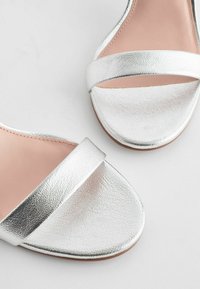 Sandales en simili cuir argenté avec deux brides parallèles, semelles plates et texture lisse. Design à bout ouvert avec une esthétique minimaliste.
