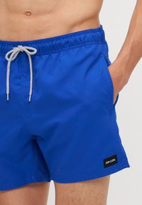 Shorts de bain bleus avec une taille élastique et un cordon blanc, dotés de poches latérales et d'un patch logo Rip Curl noir. Texture lisse.