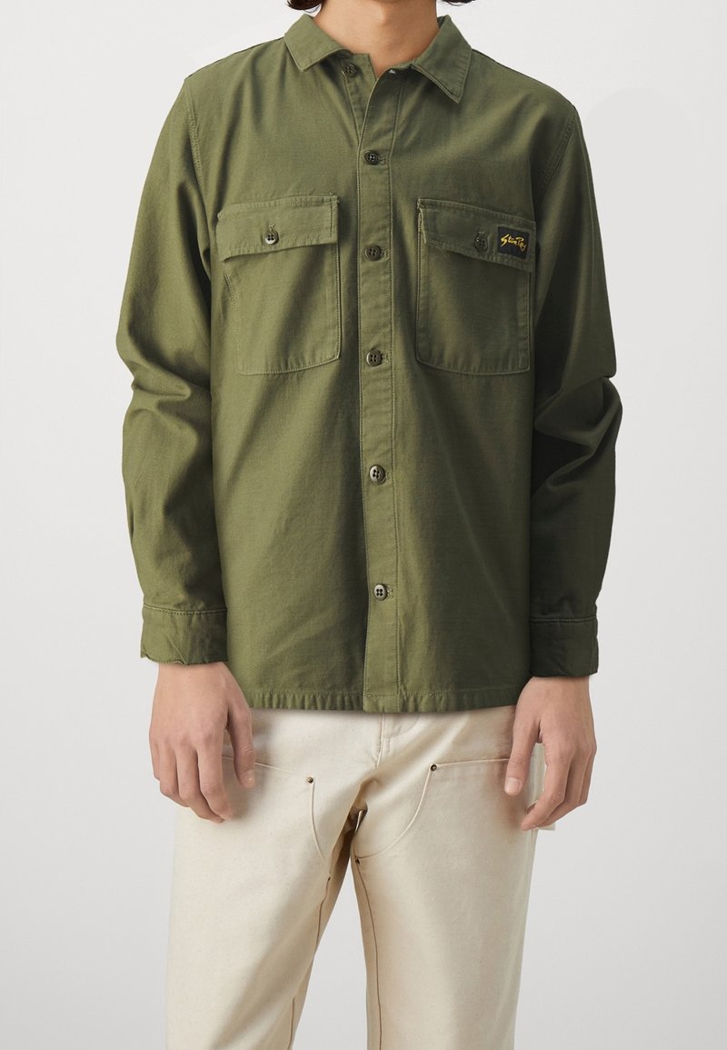 Homme portant une chemise boutonnée vert olive avec deux poches à rabat à l'avant et un pantalon crème, debout devant un fond clair uni.