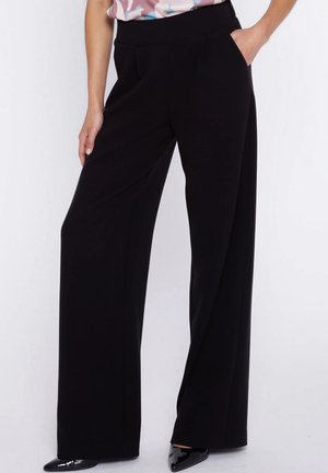Kobieta nosząca czarne szerokie spodnie z wysokim stanem i kieszeniami po bokach, w połączeniu z czarnymi lakierowanymi butami z ostrym noskiem.
