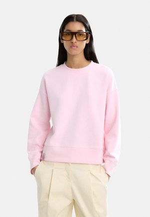 Młoda kobieta z długimi ciemnymi włosami, nosząca duże prostokątne okulary przeciwsłoneczne w brązowym odcieniu, jasnoróżowy sweatshirt i beżowe spodnie, z rękami w kieszeniach.