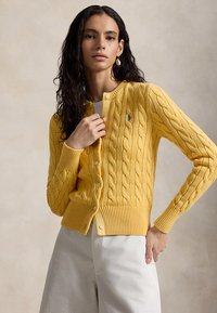 Femeie purtând un cardigan galben cu model împletit cu nasturi și pantaloni albi, pozând cu o mână aproape de guler și cealaltă pe șold.