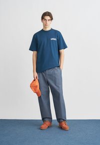 Μπλε σκούρο μπλουζάκι με λογότυπο εταιρείας, γκρι παντελόνι με φαρδιά μπατζάκια, πορτοκαλί καπέλο που κρατιέται στο χέρι και πορτοκαλί παπούτσια με κορδόνια και υφή στη σόλα.