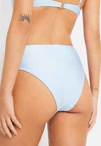 Šviesiai mėlyna aukšta bikini kelnaitė su lygia tekstūra, be siūlių krašteliais ir minimalizmo stiliumi. Pasižymi subtiliu blizgesiu ir jokiomis matomomis rašmenimis.