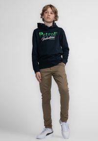 Marineblauwe hoodie met groene en witte tekst, gecombineerd met beige slim-fit broek en witte sneakers met zwarte accenten.