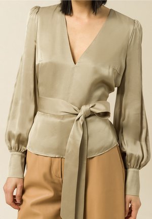 Blusa de satén en beige claro con escote en V profundo, mangas largas abullonadas y detalle de cintura anudada. Textura suave con un brillo sutil.