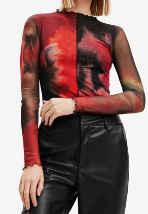 Vrouw draagt een rood-zwart tie-dye mesh top met lange mouwen en een kroezelzoom, in combinatie met zwarte, hooggesneden nepleren broek.