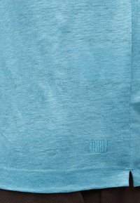 Φωτεινό γαλάζιο βαμβακερό ύφασμα με διακριτική υφή και μικρό κεντημένο λογότυπο κοντά στο κάτω τελείωμα πάνω από μια πλαϊνή σχισμή.