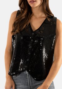 Chaleco negro con lentejuelas, con escote en V, frente de siete botones y diseño sin mangas. Textura suave con detalles brillantes, combinado con jeans grises.