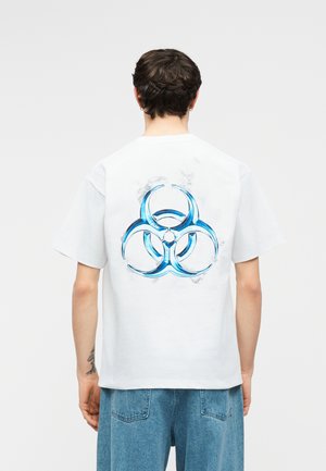 Persoon met kort donker haar draagt een wit t-shirt met een groot blauw biohazard-symbool op de achterkant en losse blauwe spijkerbroek.
