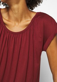 Burgundy blus med rund halsringning, veckad frontdesign och korta ärmar. Mjukt bomullsmaterial, slät textur, minimalistisk stil.