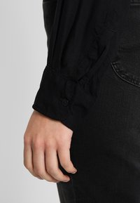 Black long-sleeve shirt with buttoned cuff, featuring soft fabric and pleated design at the arm. Worn with dark jeans.